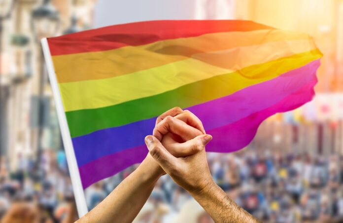 couple holding hands and wave in front of a rainbow flag